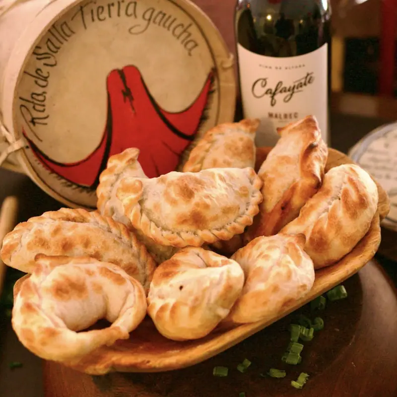 Empanadas salteñas recién horneadas en una fuente de madera, junto a un vino Malbec y un bombo legüero